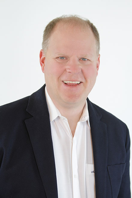 http://Head-and-shoulders%20portrait%20of%20a%20smiling%20man%20in%20a%20dark%20blazer%20and%20white%20shirt%20against%20a%20light%20background.